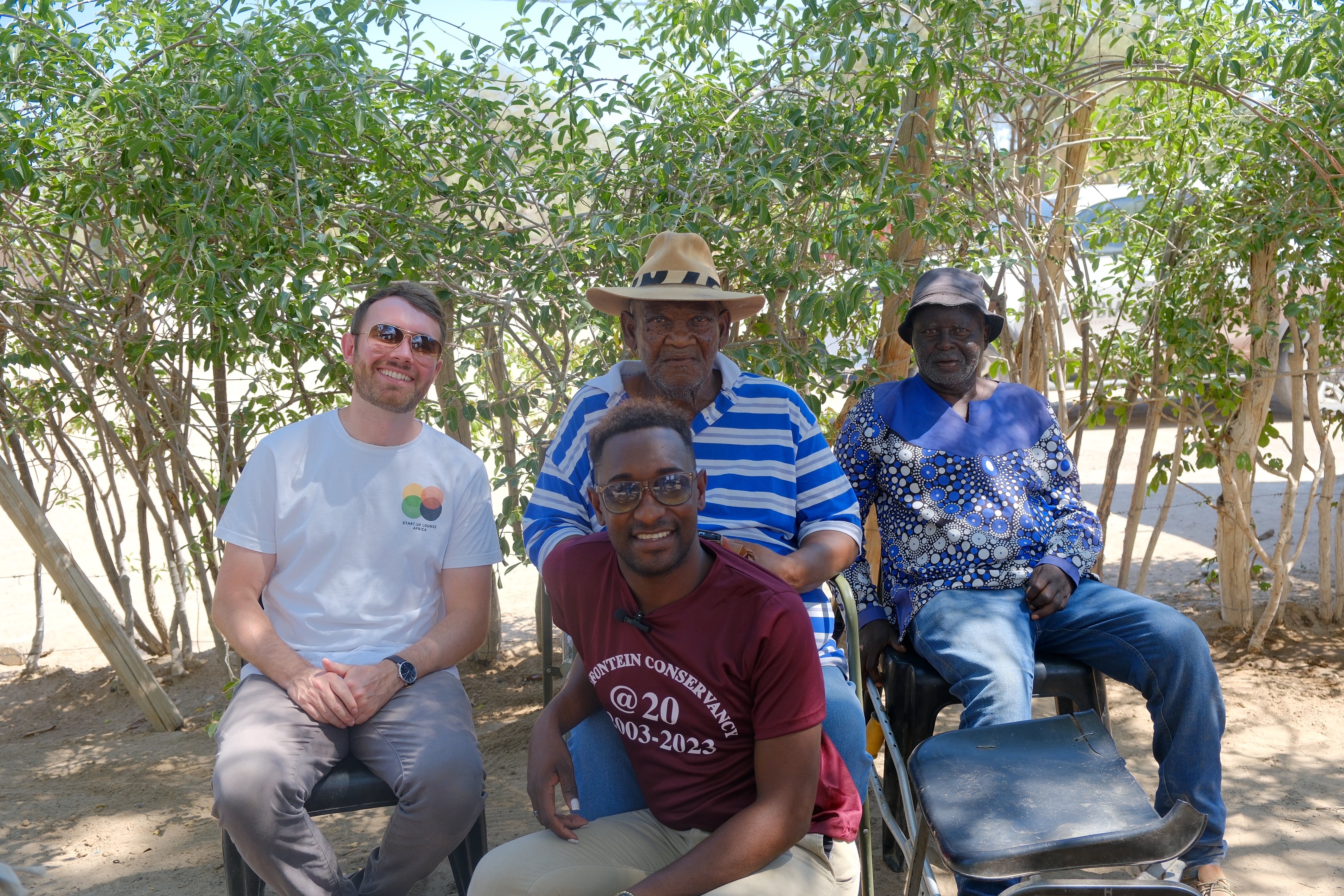 Chief Jeremiah Gaobaeb, Headman Sam Gaseb (Nami-Daman Traditional Authority) with Bentji Tourob and Nick Murphy (Our Sustainbale World), sitting in front of bushes, Namibia